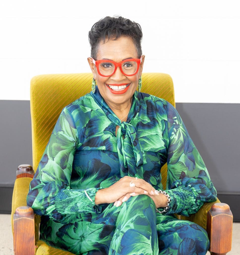 Vicki Saylor sitting in yellow chair, wearing green and blue patterned clothing, red-framed glasses and smiling