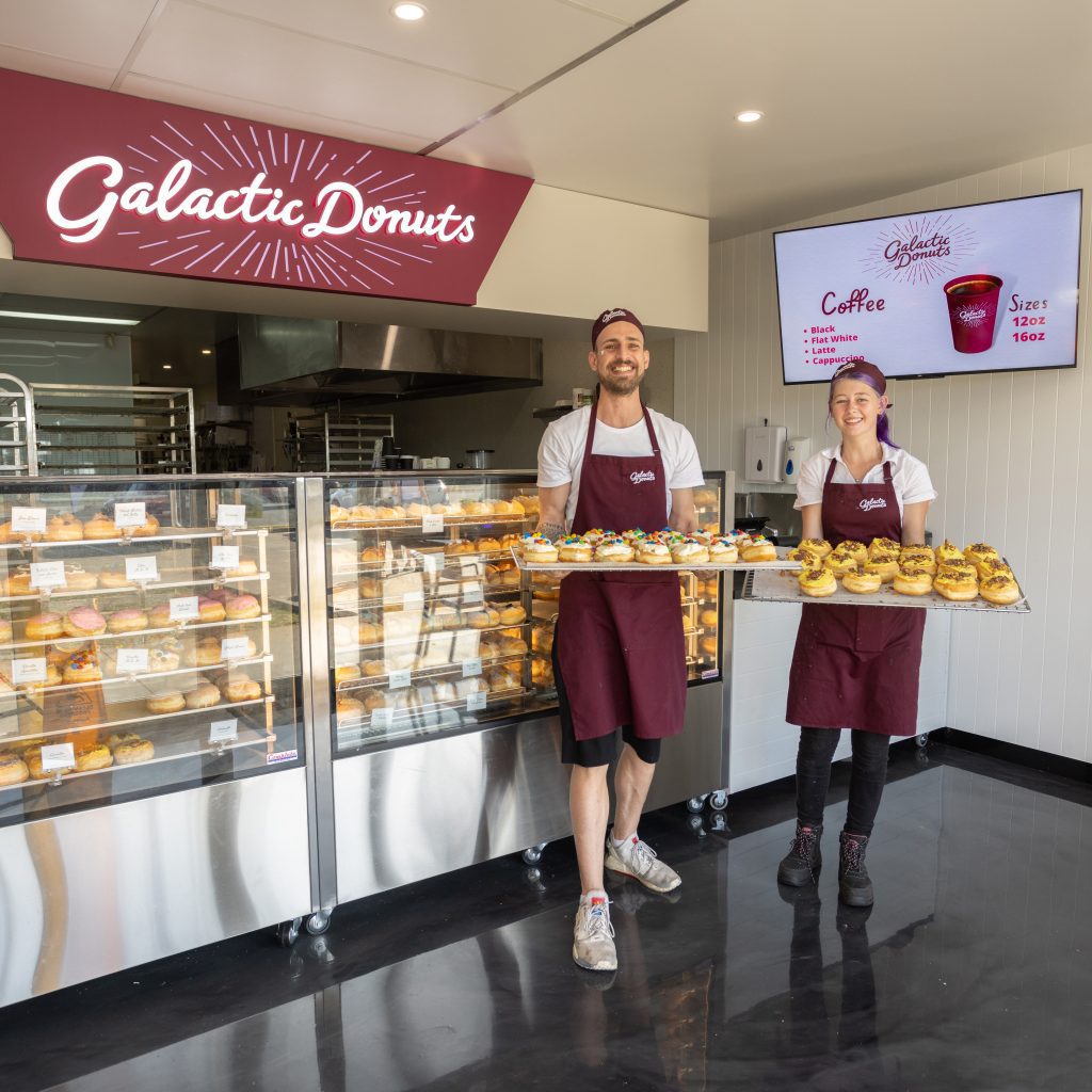 Staff members at Galactic Donuts holding trays of doughnuts in front of the shop counter and sign
