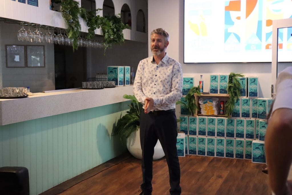 Aaron Rieniets, founder of Tan Lines Distilling, standing on front of a white and teal coloured bar. Behind him is a video screen and rows of Tan Lines branded boxes.
