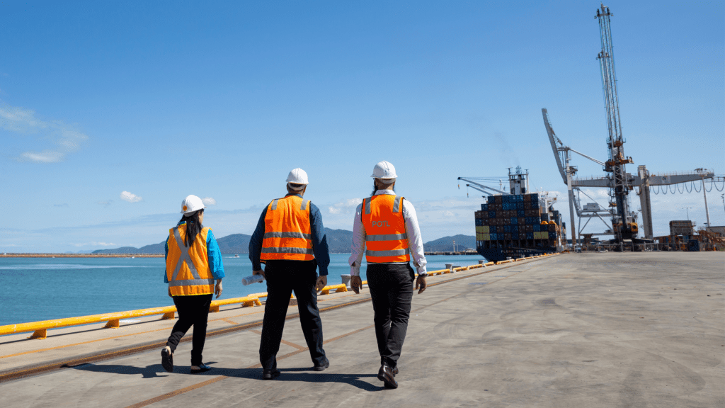Three people in hi-vis vests and hard hats walking towards container ship.