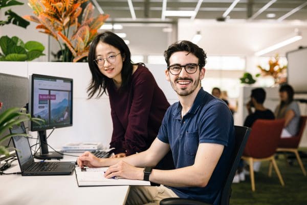 Woman standing and man sitting at desk, both looking and smiling at camera