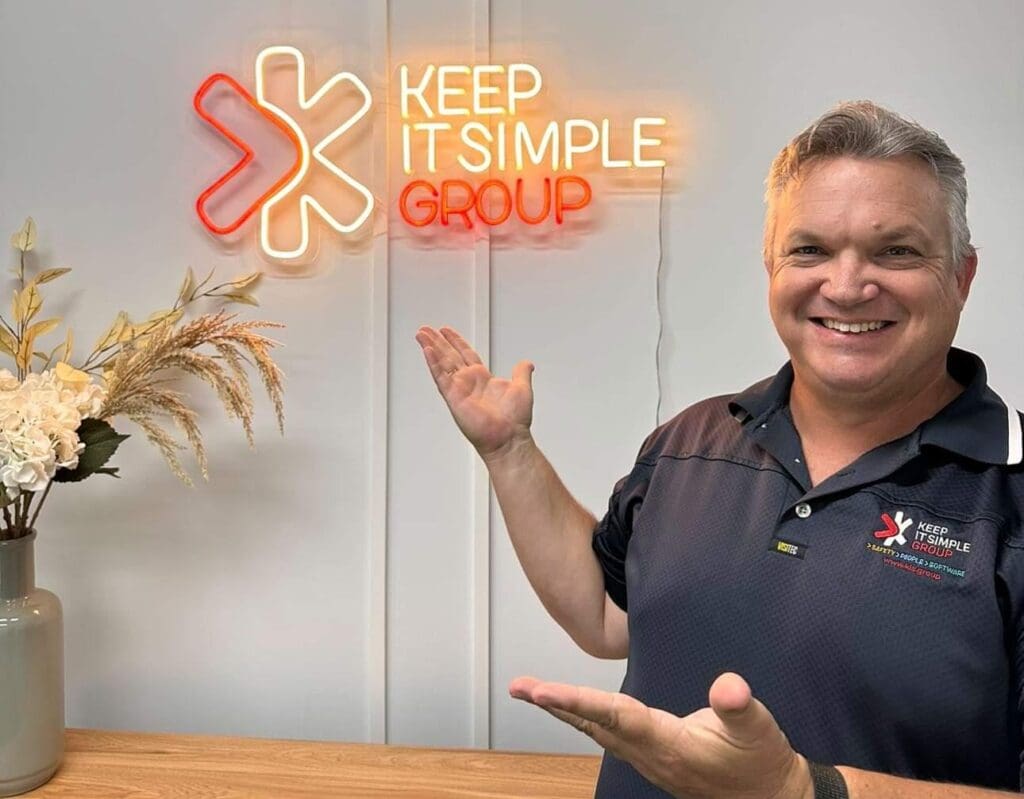 Shayne Connolly pointing at neon sign labelled "Keep It Simple Group" with vase of dried flowers also in the frame